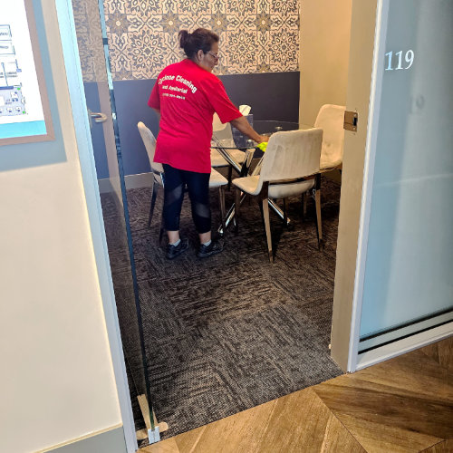 A Cyclone Cleaning and Janitorial staff member wearing a red shirt is cleaning a glass table in a modern office space, showcasing professional office cleaning services.